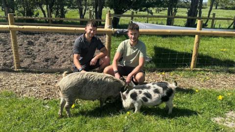 Two men with dark curly hair and dark facial hair are crouching on some grass with farm enclosures behind them. On the grass with them are two small pigs. One is a grey and beige colour and the other is black and white