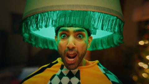 A man with his mouth open, looking shocked. He has a lampshade on his head and has on a baggy orange, black and white top
