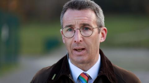 Andrew Muir has short greying hair and is wearing glasses. He is wearing a brown jacket over a white shit with a multi-coloured tie.
