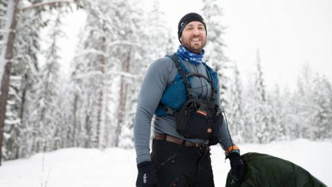 Rob Wilkinson pictured during the challenge. He is wearing a black hat and trousers, a front facing bag, blue scarf and a light blue top. Snow and fir trees are in the background of the image.