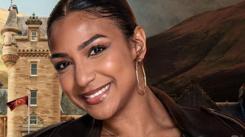 A close-up of Maia, a 25-year-old woman. Her dark hair is pulled back from her face and neck. She is tilting her head to her right and is smiling broadly. She is wearing large gold hoop hearings and a brown collared leather jacket. Behind her is a Scottish hill and a stone tower with a grey slate roof