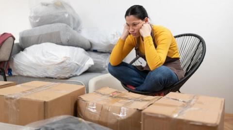 Young woman is sitting on the chair and looking at all of the boxes that need to be unpacked in her new home