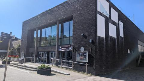 A general view of the entrance to the Civic Hall in Whitehaven. It is a one-storey building with a flat roof, built in dark bricks, with a glass-fronted entrance.