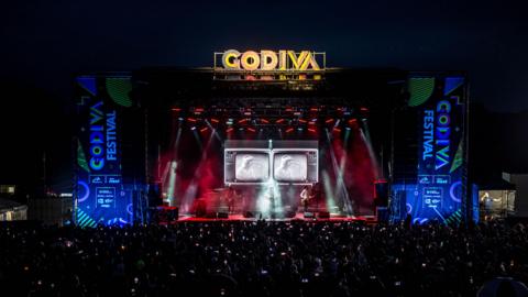 The main stage at Godiva Festival at night, with red and blue lighting around the stage, and a large Godiva sign illuminated in yellow on top of the stage. There is a large crowd silhouetted in front of the stage and, as it's dark, you can see the lights of mobile phones that people are holding in the air. 