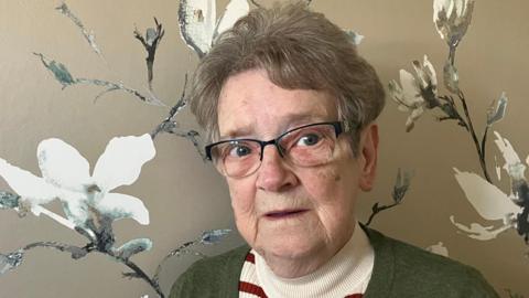 A woman in a green cardigan and wearing glasses looks at the camera.