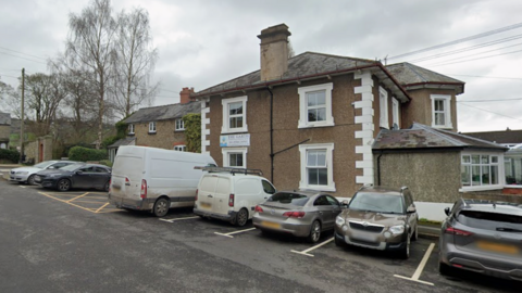 A two storey building with a brown cobbled exterior and white window pains. There are cars parked in the car park alongside it. 