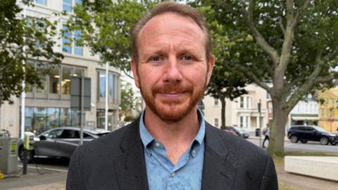 Tom Coles is wearing a black blazer and a blue shirt. He is pictured in front of the government building. 