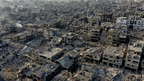 An aerial shot shows vast destruction: bombed out buildings, rubble, and rising smoke. 