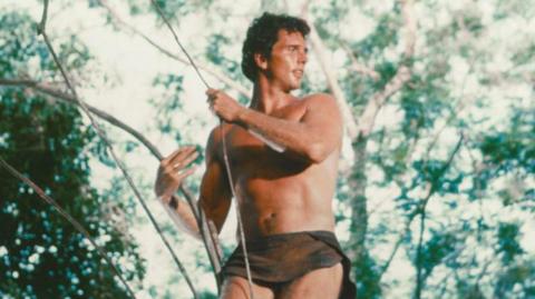 Ron Ely dressed in a loincloth, holding on to a vine in a tree