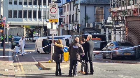 Four people, including a man with a black police jacket on, stand in front of blue and white police tape stretched across the road. Beyond the cordon is a crashed white car on its side. There is a police forensics officer in a white suit near the car.