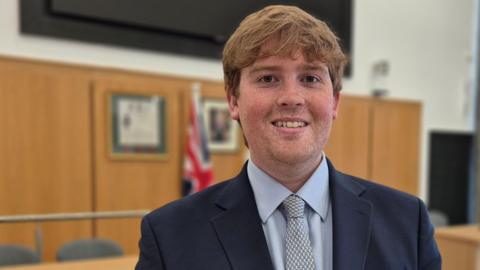 Dan Swords wears a navy blue suit, blue shirt and patterned tie. He smiles at the camera, while standing in a high-ceiling council chamber, with a large black screen on the wall. He has ruffled light brown or ginger hair.