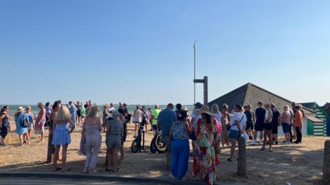 People gathering on a beach. It is a sunny evening. The sea is in the distance and roofs of huts can be seen.