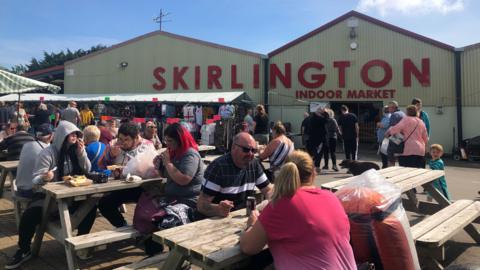 Several benches located in an outside seating area with several people sat on them with canned drinks and food. In the background is a warehouse-type building which says 'Skirlington Indoor Market' in large red letters.
