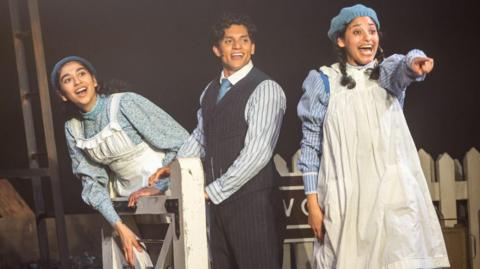 Three Railway Children actors stand on a platform and look to their left. One is pointing and all are smiling. 