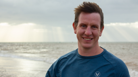 A man with brown hair and a blue tshirt smiling on a beach
