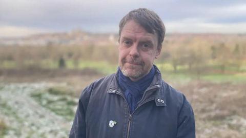 A man standing in a field. He has short brown hair and brown/grey stubble. He is wearing a navy jacket with a white flower pin badge. He has a blue scarf.