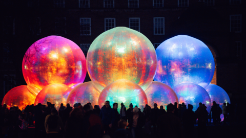 Lots of colourful bubbles layered on top of one another in the dark. There are three larger bubbles on top of seven smaller bubbles. They are lit up and there are people stood in front of them. 