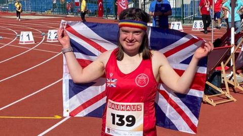 A woman smiling at the camera, hands aloft holding a union jack flag behind her back. wearing a red athletic vest and standing on an athletics track. She's also wearing a rainbow coloured sweat band on her head.
