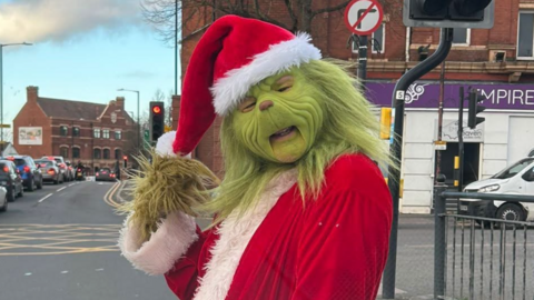 A man dressed up as the Grinch standing at a crossing. Cars are queuing in traffic in the background. 