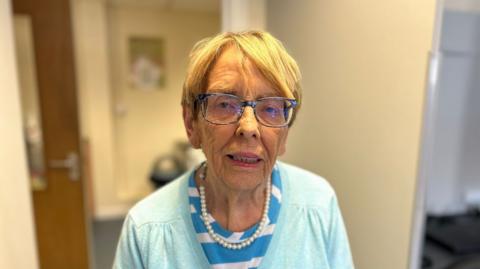 A woman with short light coloured hair is looking into the camera. She is wearing dark rimmed glasses, a blue and white striped top and a light blue cardigan with a pearl necklace. 
