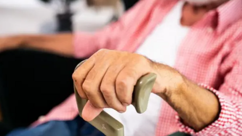 An elderly man places his left hand on a walking stick while seated. He has a pink shirt on with a T'shirt on underneath.