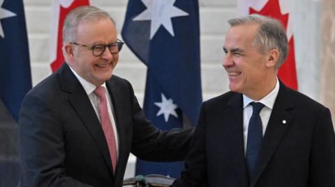 Two middle aged men with grey hair and wearing dark suits smile in front of the Australian and Canadian flags.
