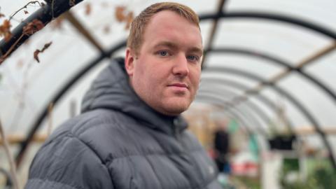 Josh Andrews, who has short red hair combed to the left, stares directly at the camera with a serious expression on his face. He is standing in a dome-like greenhouse which is blurred. Josh is wearing a grey down-filled hooded jacket.