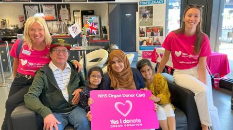 From left to right, Lisa Wilson with recipient Gordon, recipient Fatima and her mother Lubna and sister Sofia, and her daughter Pippa. They are sitting on a sofa, smiling for the camera. The mother and two daughters are holding a pink NHS Organ Donor Card with a heart in the middle and the message "Yes I donate". Various Transplant Games merchandise is displayed behind them, as well as information about Lisa's son, Tom.