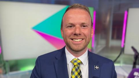 Ben Maguire is smiling broadly at the camera. He is wearing a white shirt, blue jacket and a Cornish tartan tie. He has short blond hair and a slight beard. There is  St Piran flag badge in the button hole of his jacket. Behind him are neon-lit panels.