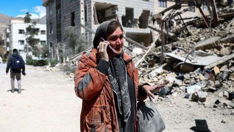 A woman talks on a phone while standing amid a damaged residential neighbourhood hit by a strike as the U.S.-Israeli conflict with Iran continues, in Tehran, Iran, March 30, 2026.