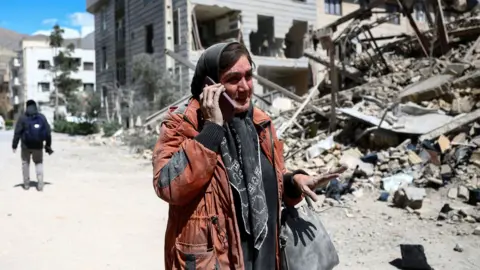 A woman talks on a phone while standing amid a damaged residential neighbourhood hit by a strike as the U.S.-Israeli conflict with Iran continues, in Tehran, Iran, March 30, 2026. 