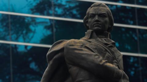 A large, black statue of a man dressed in late-18th century clothes, whose hand is on a sword by his side. The backdrop is a large office building with mirrored glass windows which reflect the blue sky.