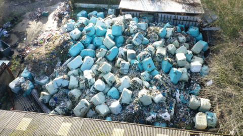 An aerial view of bales of rubbish which have been dumped on a farm in Lincolnshire. The blue-coloured bundles are piled up and surrounded by two agricultural buildings.