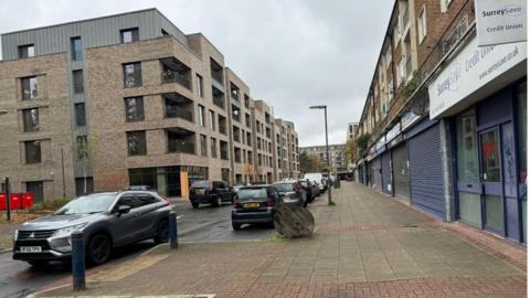 Dartmouth Avenue in Sheerwater. On one side of the road are the new flats which have been built. On the other side are older buildings with shops underneath them. One of them has a sign for SurreySave Credit Union.