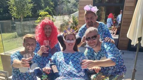 Found men holding wine glasses and a woman in the middle. They are all wearing mermaid tail inspires tops that are a light blue with scales. They are wearing party accessories and smiling at the camera. They are outside. 