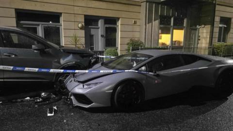 A silver Lamborghini behind police tape after crashing into a Kia.