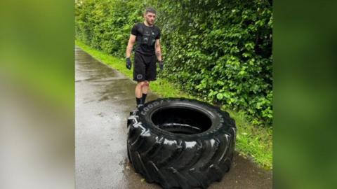 Will Fraser-Coombe, wearing black sports top, shorts and gloves, readying himself to flip a 100kg tyre. The path is wet and it is clear it has rained. 