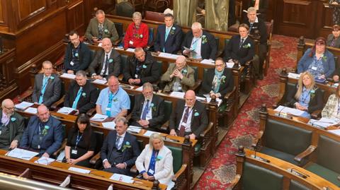 A bird's eye view of a council chamber, focused on several rows of seats featuring people wearing turquoise rosettes, identifying them Reform UK councillors