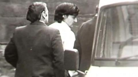 A black and white still from TV footage showing Peter Sullivan, who has thick black hair and is wearing a white shirt, being led to a police van in handcuffs.