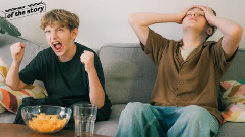 Two boys watching football match, one celebrating, one miserable