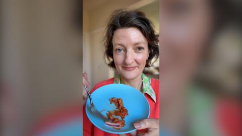 A woman with brown hair holds up a blue plate holding kimchi. She is wearing a dark pink jumper and a green-collared shirt.