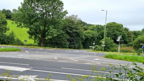 A road junction on a dual carriageway. There are green trees in the background. The sky is grey. 