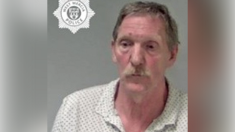 Mug shot of a white man with short hair and a moustache. He is wearing a patterned white polo shirt.