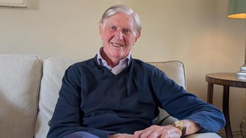 Bill Cash wears a navy V-neck jumper over an open button collar shirt. He is smiling at the camera. He is sat on a sofa next to a table with a lamp on it. 