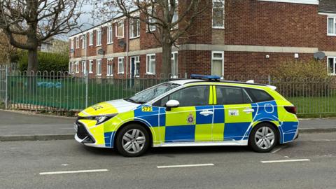 A police car parked up by a residential block of flats. The car is yellow, blue and white. The flats behind are red brick and there are many white framed windows.