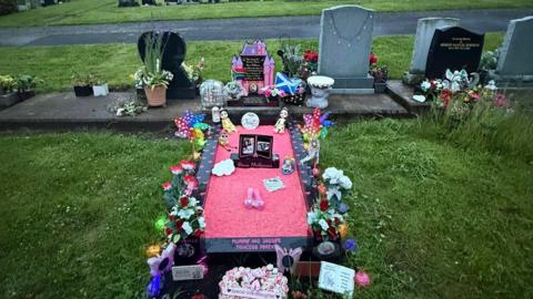 A graveyard, and the central grave is decorated with pink gravel, a black stone border and decorations around the grave that are representative of a child. the headstone is a Disney castle with a South African and Scottish flag on both sides.