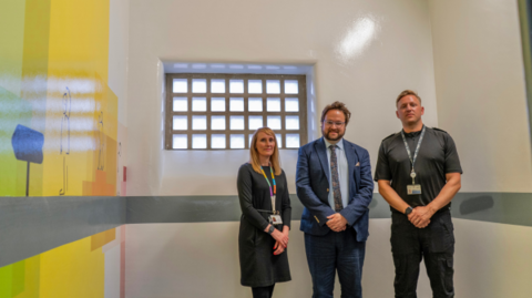 Three people stand in a small white room facing the camera. The new "youth focussed custody suite" has a colourful painting on one wall. The small window at the top of the room has a criss-crossed bar patterned frame.