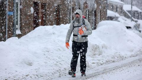 A man in an grey anorak walks on a snowy road with large banks of snow at the side of the road