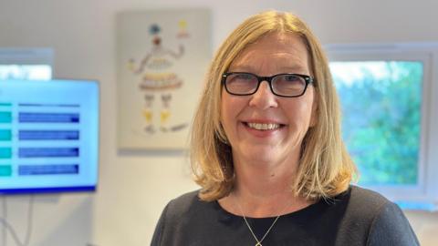 A head-and-shoulders picture of Gill Haigh, managing director of Cumbria Tourism. She has blonde shoulder-length hair and blue eyes. She wears glasses with black rim, a dark top and silver necklace.