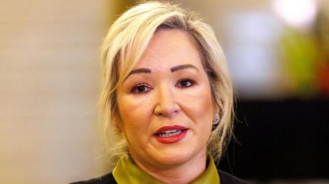 Michelle O'Neill. She has shoulder-length blonde hair, wearing glasses, and a green blouse.